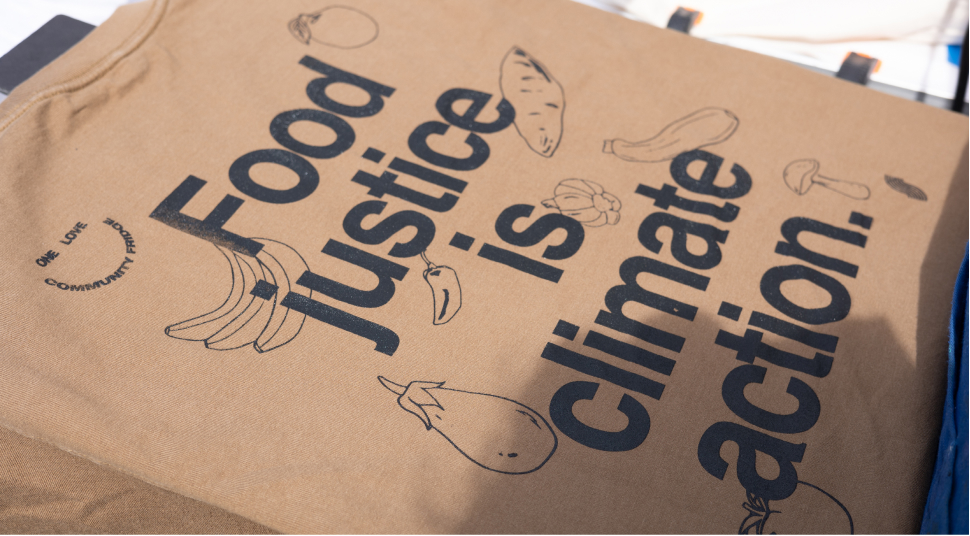 A brown screen printed t-shirt with illustrations of produce that reads, “Food justice is climate action.”