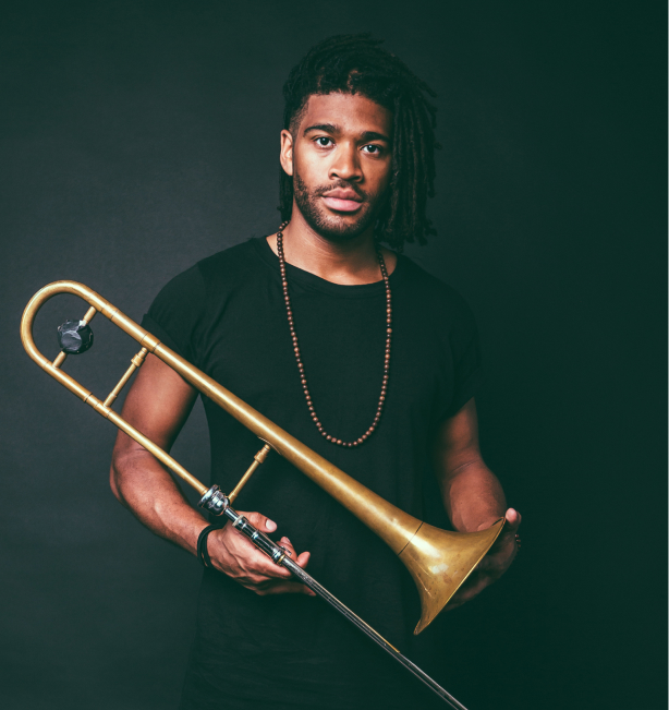 Darius Christian, a black man with chin-length braids and facial hair holds a trombone. He is wearing all black and stands in front of an all black backdrop.